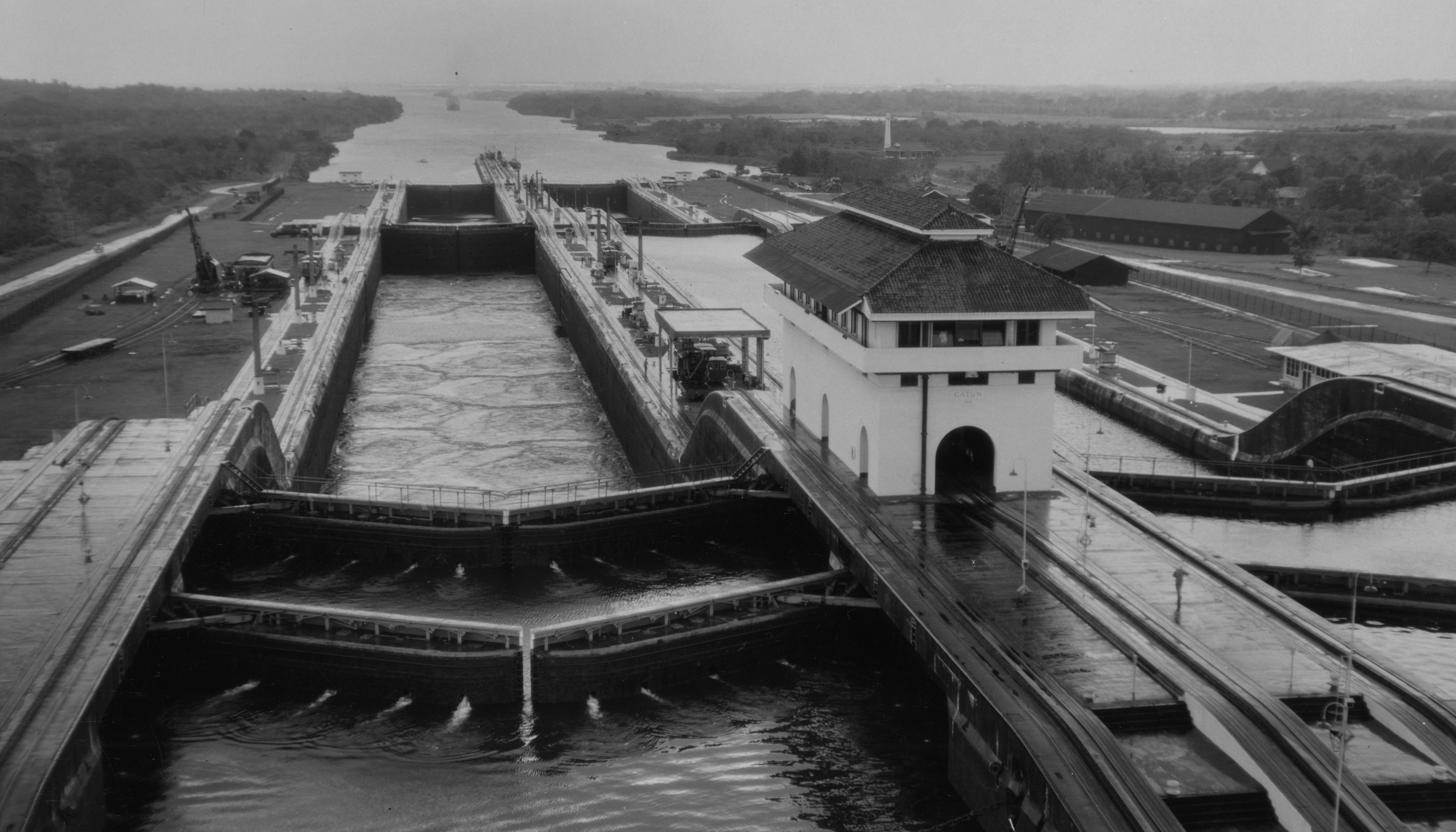 Černobílý, zvýšený pohled na Gatun zámky na Panamském kanálu. Masivní komory zámku betonu jsou naplněny vodou a na pravé straně zámků stojí velká bílá kontrolní budova s tmavou střechou. Přes branky procházejí dlouhé úzké mosty. V dálce se rozprostírá velká část vody, pravděpodobně jezero nebo jiná část kanálu, směrem k obzoru, ohraničené zalesněnou zemí. Podél okrajů zámků jsou viditelné různé struktury a zařízení.