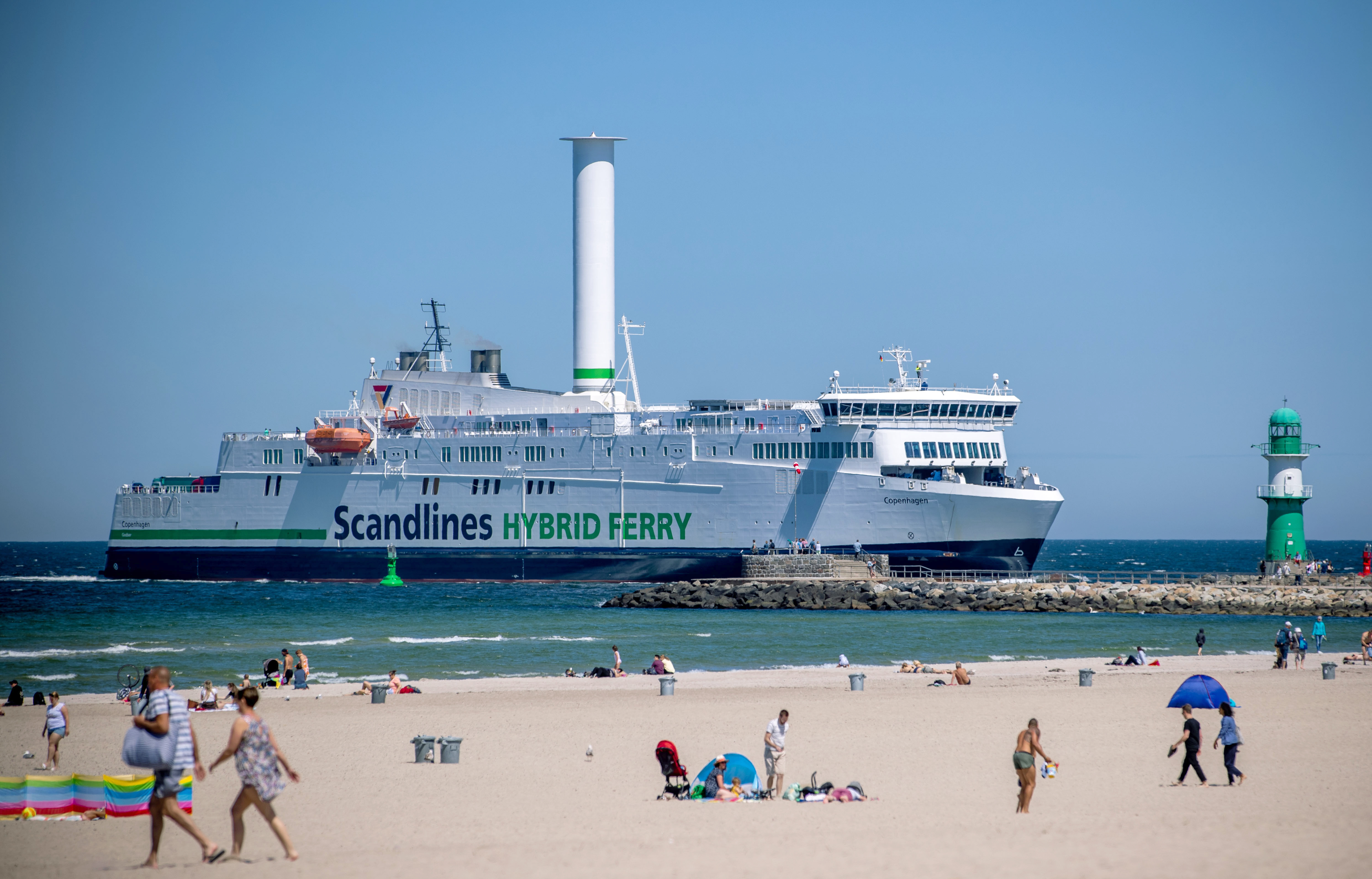 Barevná fotografie ukazuje velký bílý hybridní trajekt "Skandliny" a "Hybridní trajekt" napsáno na jeho boku. Trajekt má na palubě vysokou, bílou, točící se rotorovou plachtu. Pohybuje se přes modré moře poblíž písečné pláže s lidmi opalováním a chůzí a v dálce je viditelný zelený maják. Obloha je jasná a modrá.