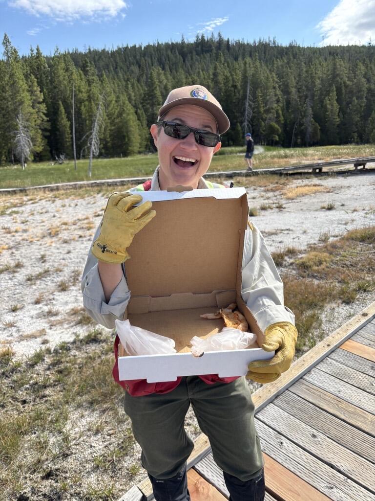 Yellowstone National Park Geology Program Dobrovolník Tara Cross se usmívá a předvádí pizzu nalezenou na Geyser Hill v Upper Geyser Basin. Kredit: National Park Service Photo by Margery Price, srpen 2025.