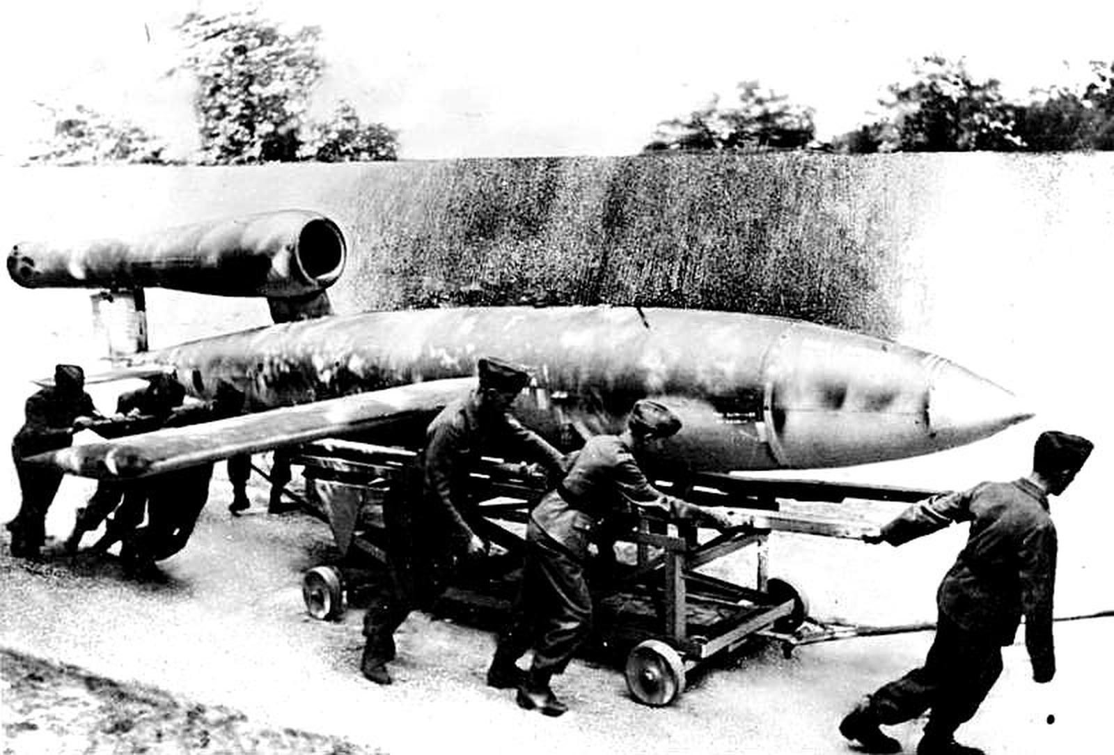 Černobílá historická fotografie ukazuje několik mužů v německých vojenských uniformách, které tlačí velkou dlouhou létající bombu V-1 (pulzně poháněná raketa) na malém kolovém vozíku. Raketa má vzadu viditelná krátká křídla a vstup motoru. Scéna se odehrává venku na zpevněné oblasti se stromy a štětcem na pozadí.
