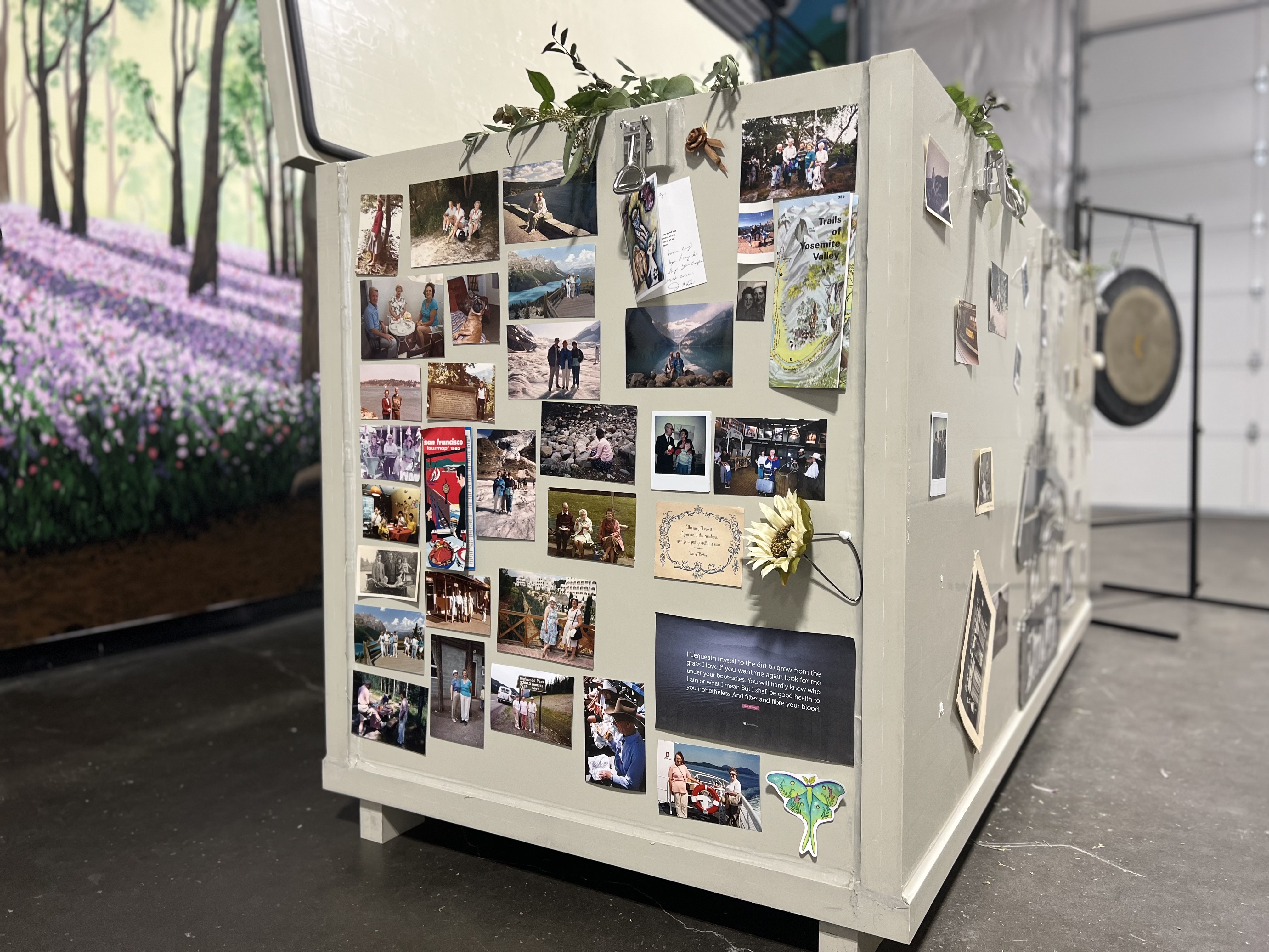 Fotografie velké, světle šedé, obdélníkové kompostovací nádoby. Strana nádoby je zdobena desítkami osobních fotografií, pohlednic a malých mementos, připojených s magnety. Na pozadí vlevo je barevná nástěnná malba zobrazující les s fialovými květinami a na stánku na pravé straně je viditelný velký kovový gong. Podlaha je tmavý beton.